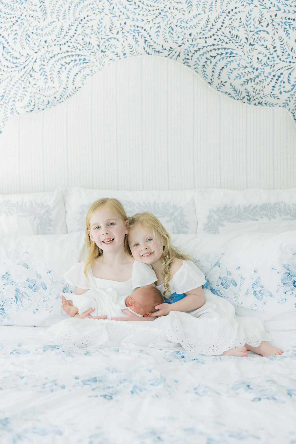 Two young sisters sit on a bed while holding their newborn baby sibling together the red balloon summit nj