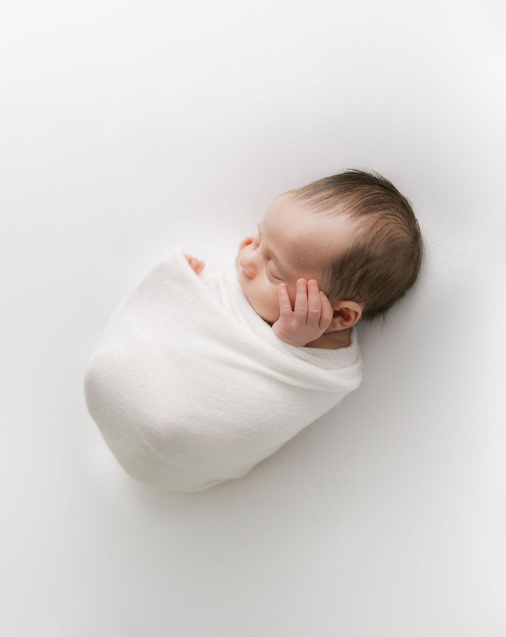 A newborn baby sleeps in a white swaddle with hands sticking out