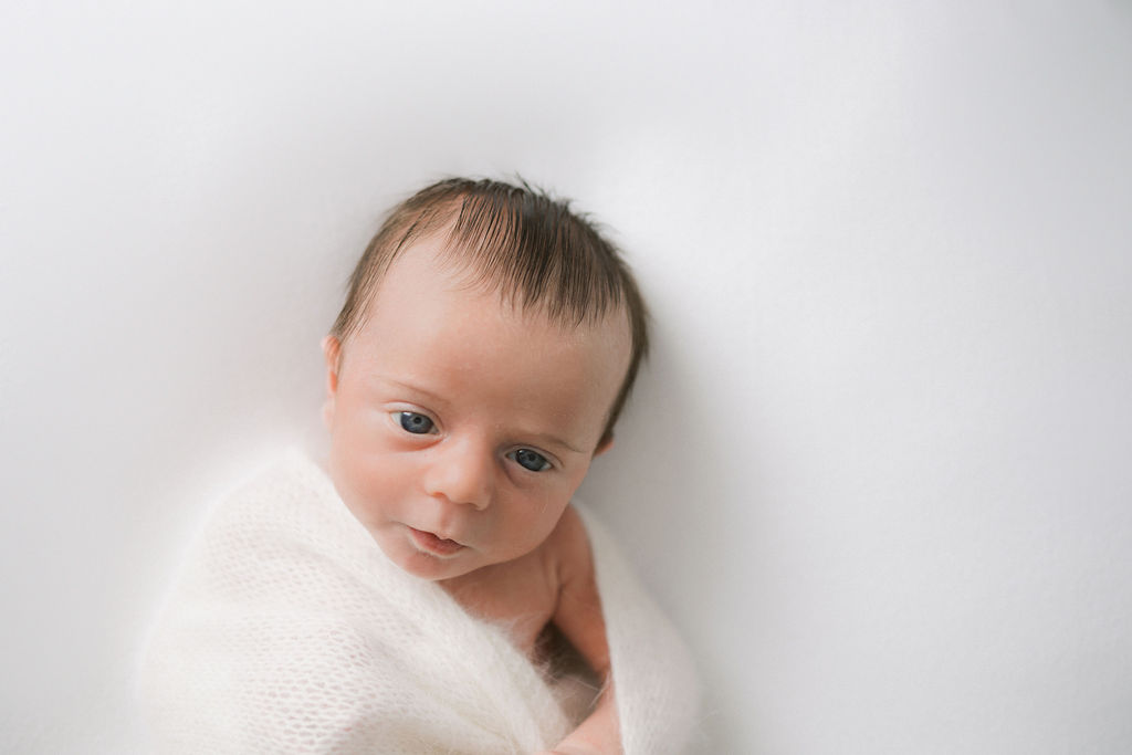 A newborn baby opens its eyes while laying on a white bed in a knit blanket baby swim lessons nj