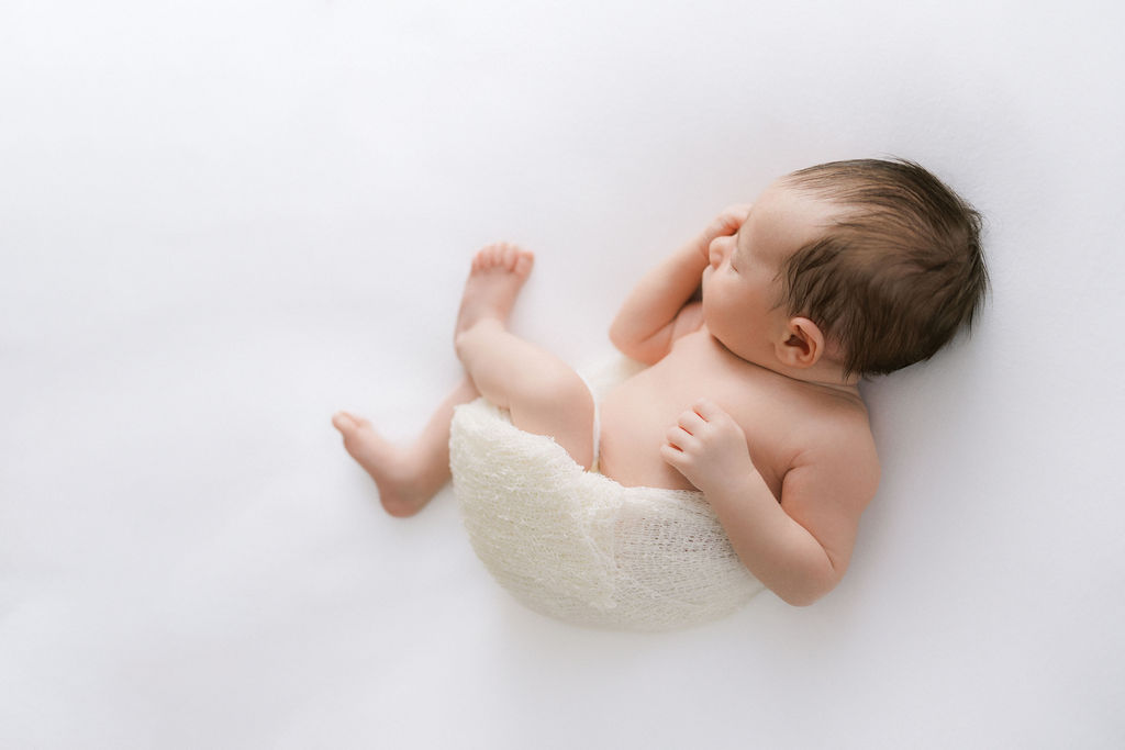 A newborn baby sleeps in a cheesecloth swaddle on a white bed in a studio baby swim lessons nj