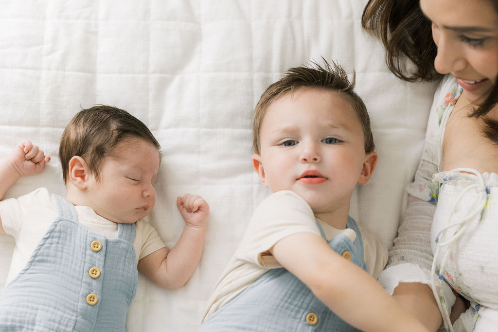 A young boy lays on a bed with his newborn baby brother in a matching blue overall outfit with mom at his side boystory boutique