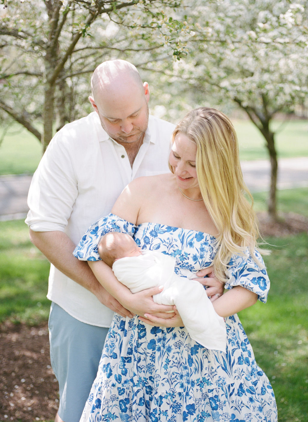 A new dad hugs mom while she holds their sleeping newborn baby and stands in a park under flowering trees