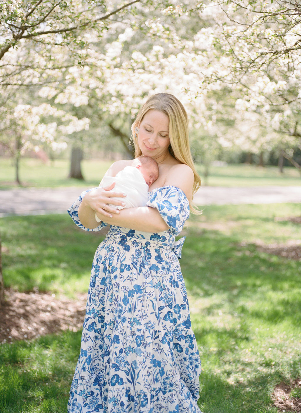 A new mom cradles her sleeping newborn against her chest while standing in a park under white blooming trees hazel jersey city