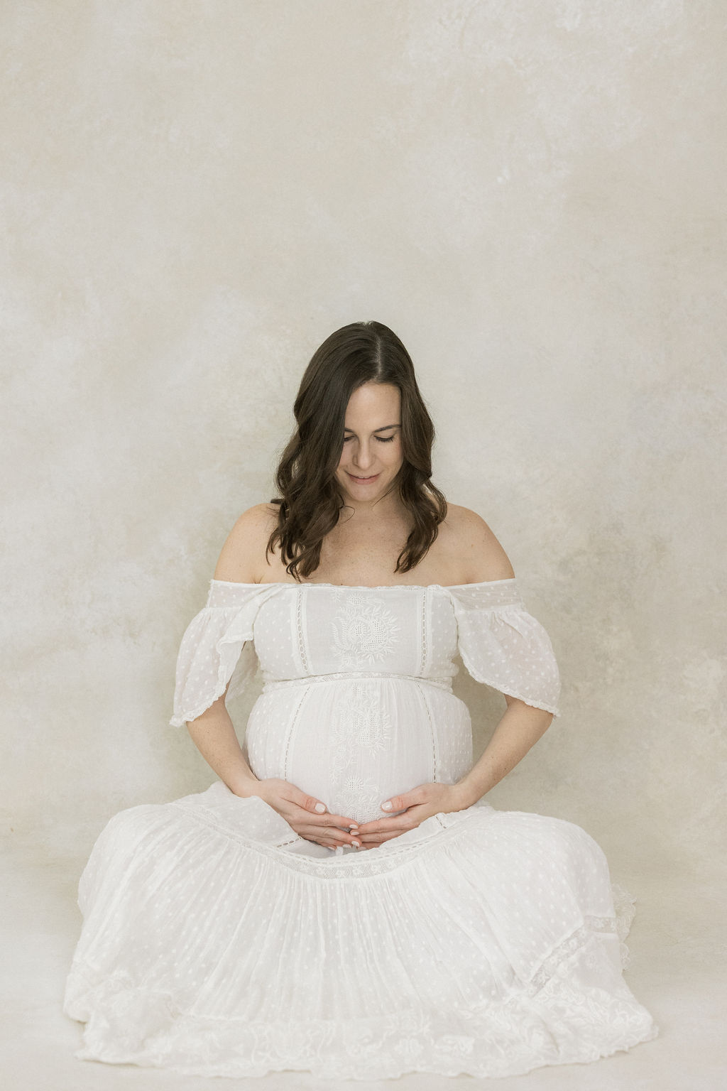 A mom to be sits on the floor of a studio smiling down at her bump with hands under it
