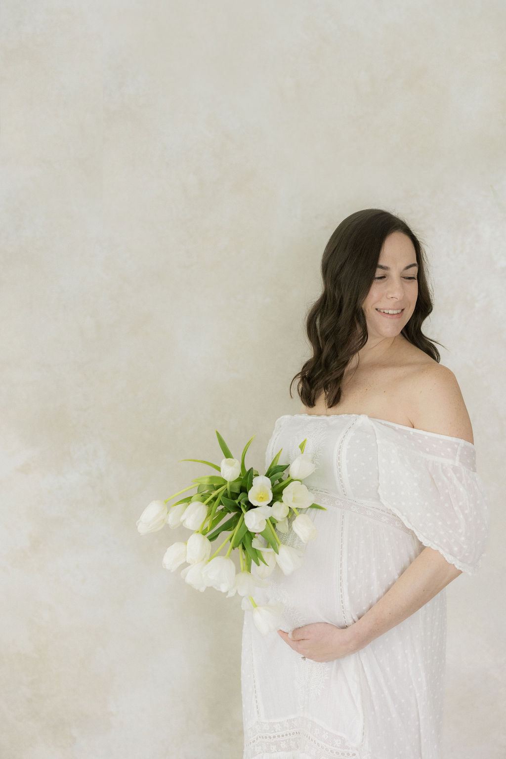 A mom to be stands in a studio holding a bouquet of white tulips in a white maternity gown nj baby shower venues