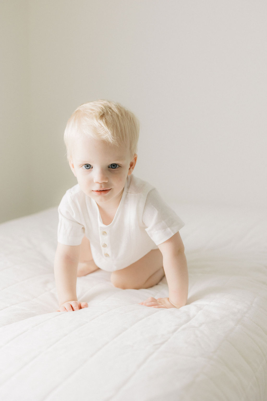 A young boy in a white onesie crawls across a white bed in a studio