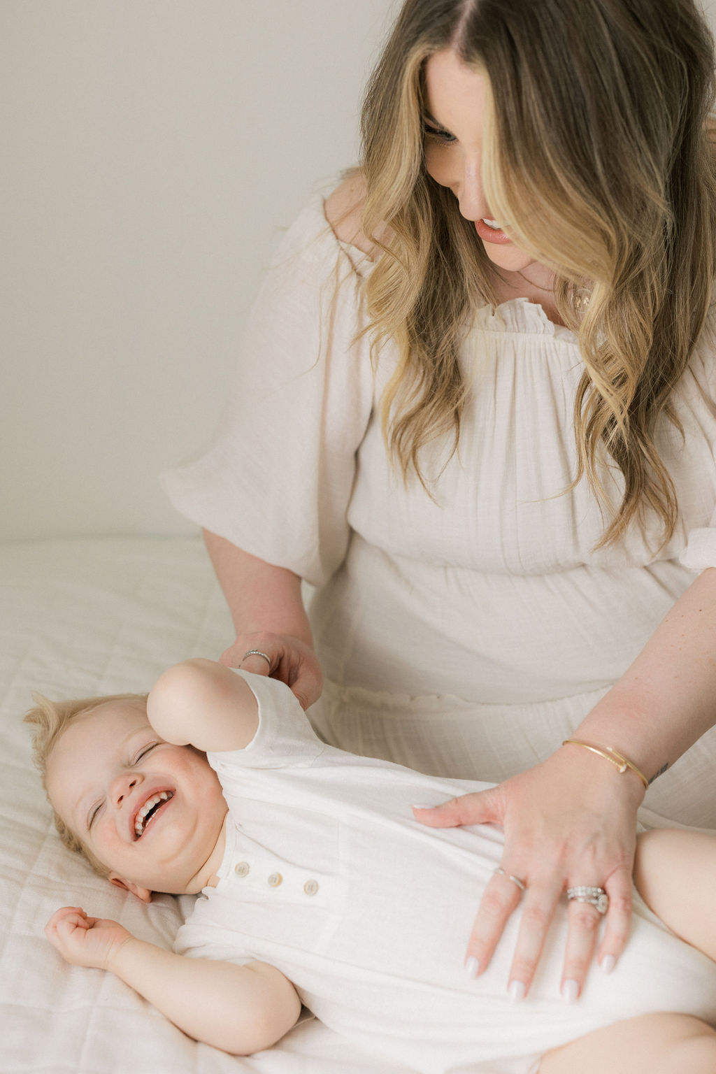 A mom sits on a bed in a studio tickling her toddler child in a white onesie nj baby stores