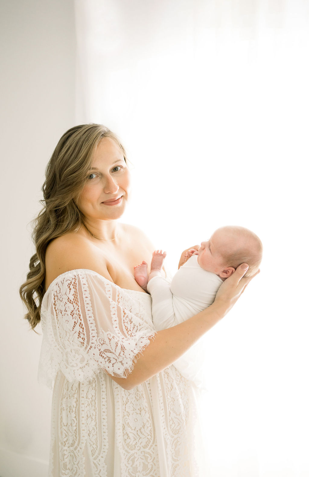 A new mom stands in a window in a studio wearing a white lace gown with her newborn baby in her arms nj birth center