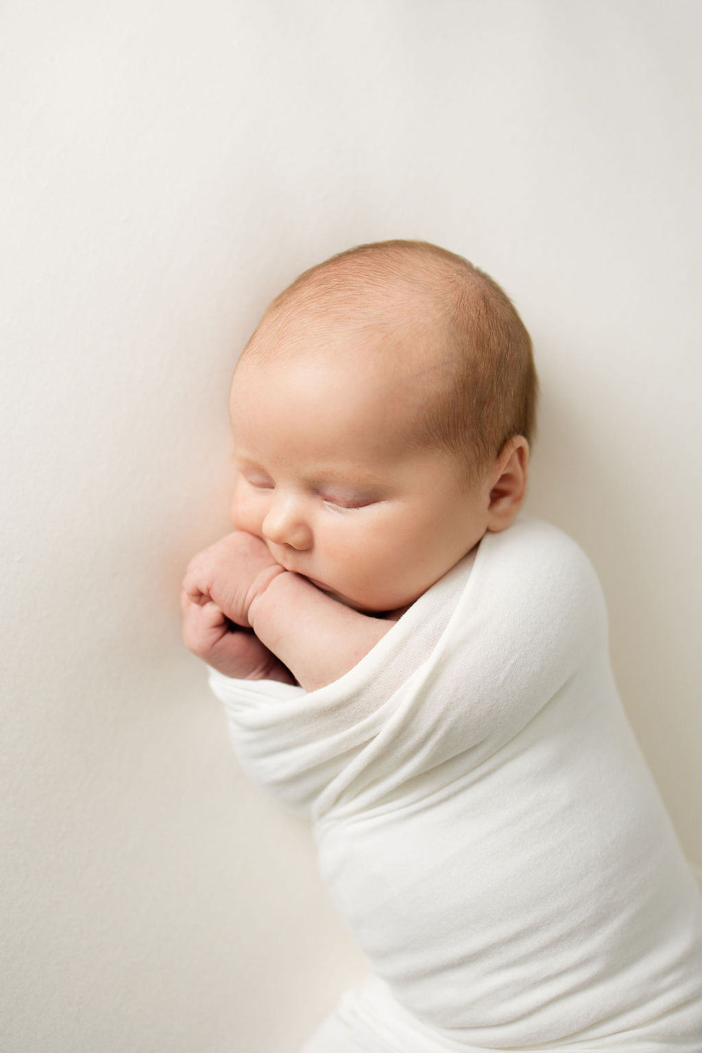 A newborn baby sleeps in a tight white swaddle with hands out