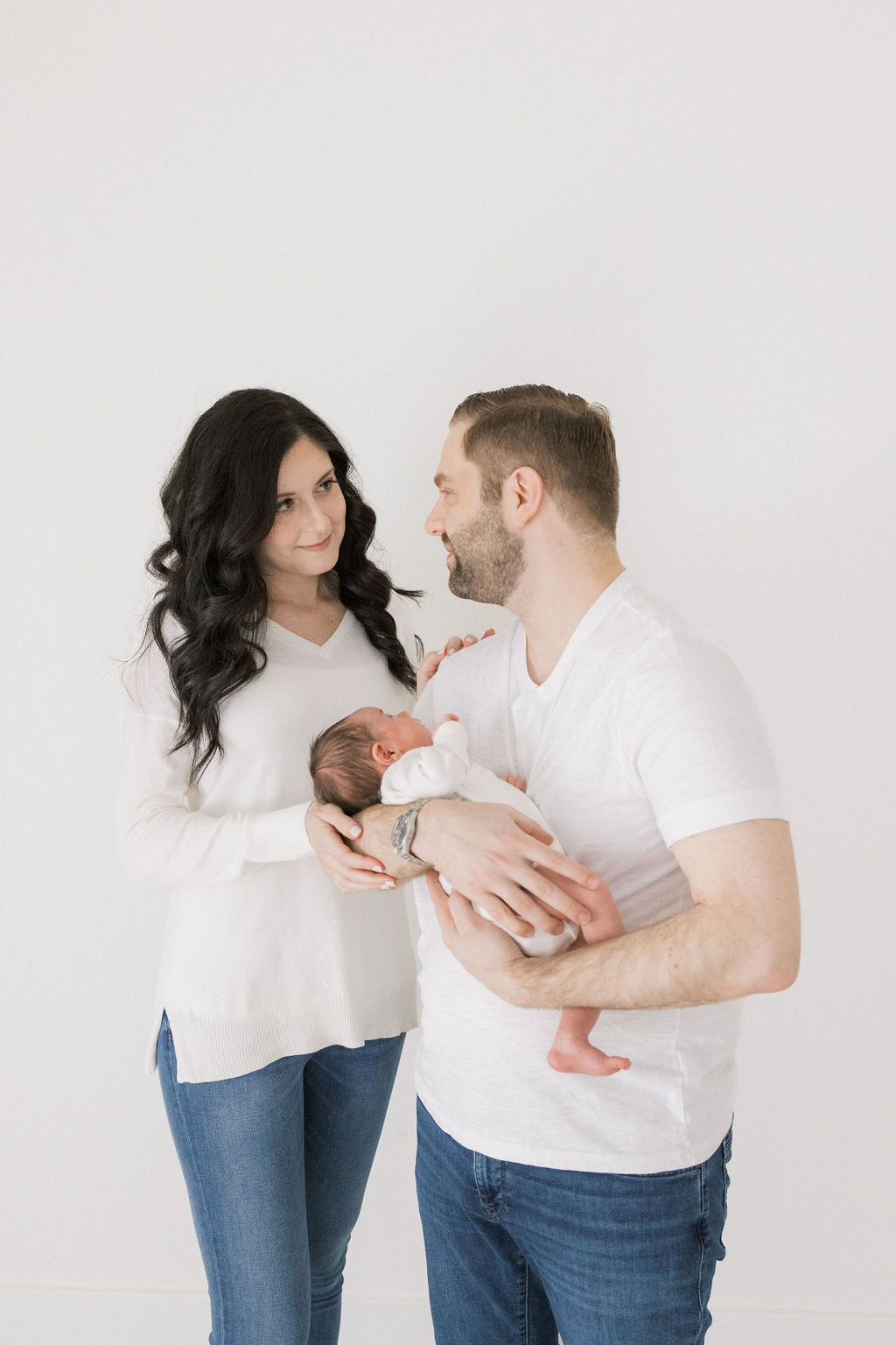 New parents smile at each other while standing in a studio holding their newborn baby after visiting bonne nuit new york