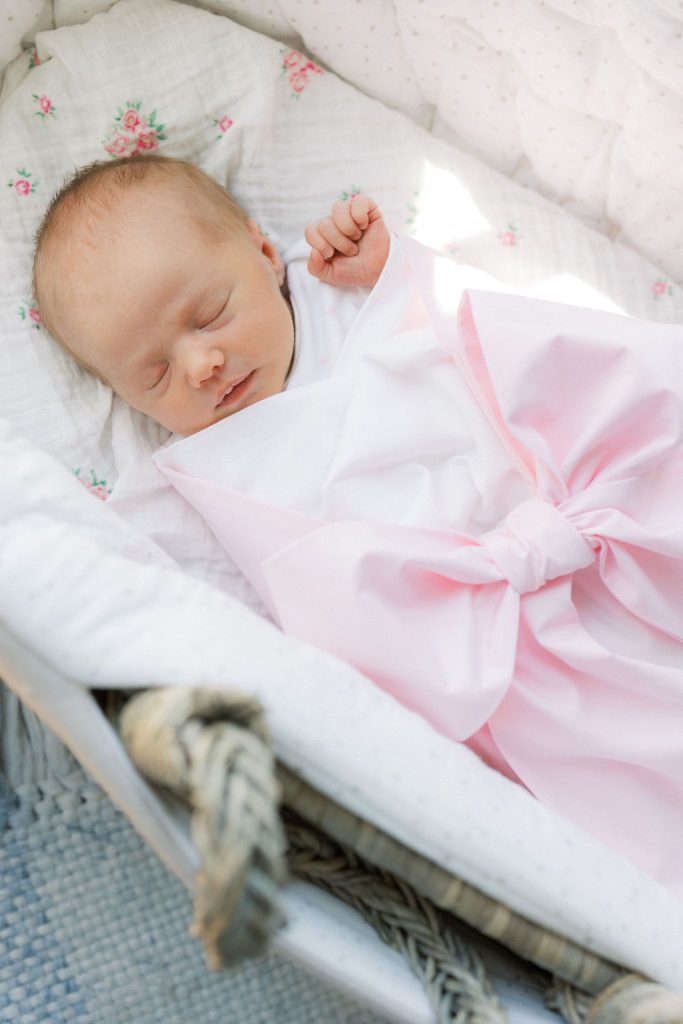 A newborn baby sleeps in a wicker crib under a large pink bow after using car seat installation new jersey