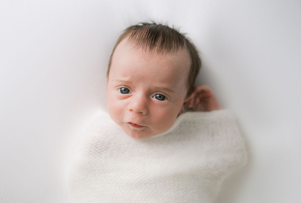 A newborn baby lays on a white bed in a knit swaddle with eyes looking straight up