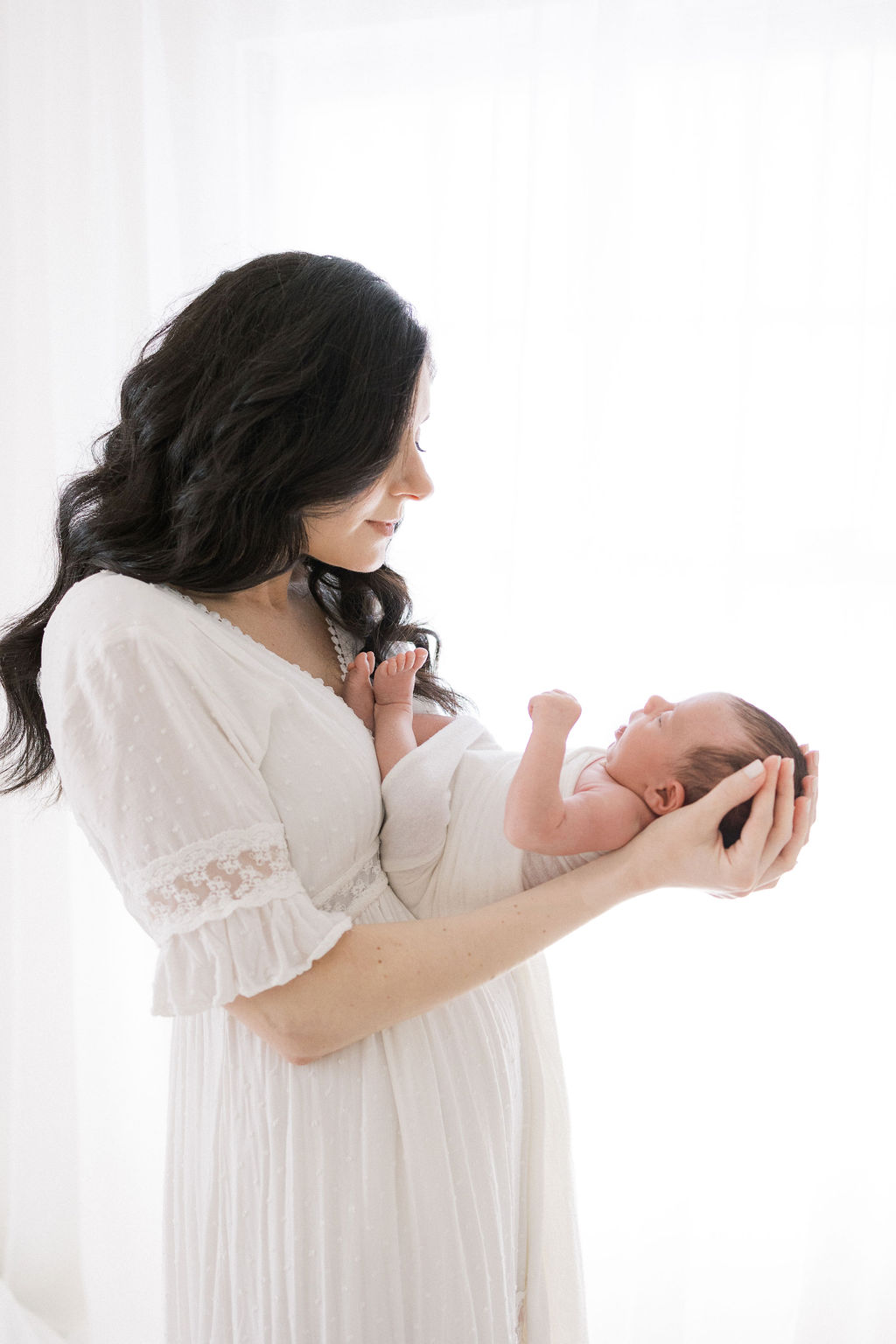 A new mom cradles her newborn baby in her hands while standing in a window before visiting cutie pie baby