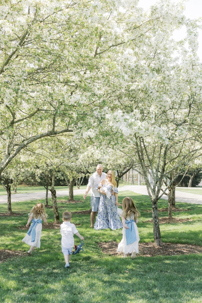 Three toddler sibling run towards mom and dad standing under white flowering trees in a park after visiting daycare in madison nj