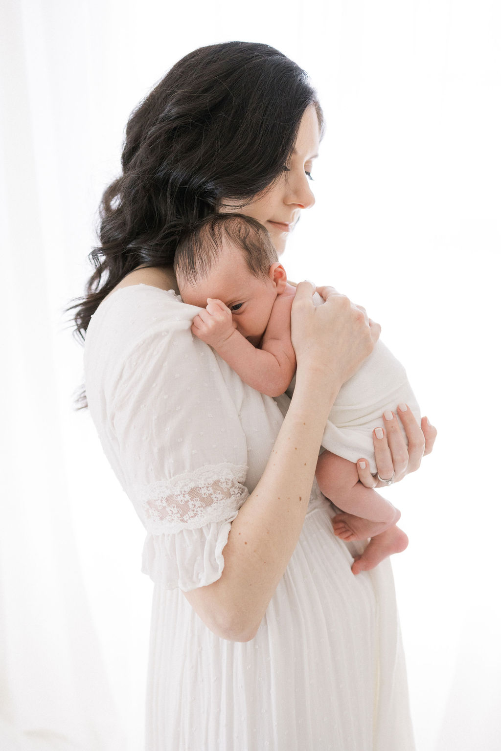 A new mom stands in a window cradling her newborn baby on her shoulder in a white dress thanks to holistic obgyn nj