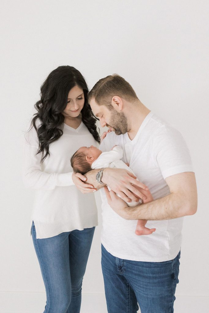 New parents smile down at their newborn baby in dad's arms while standing in a studio