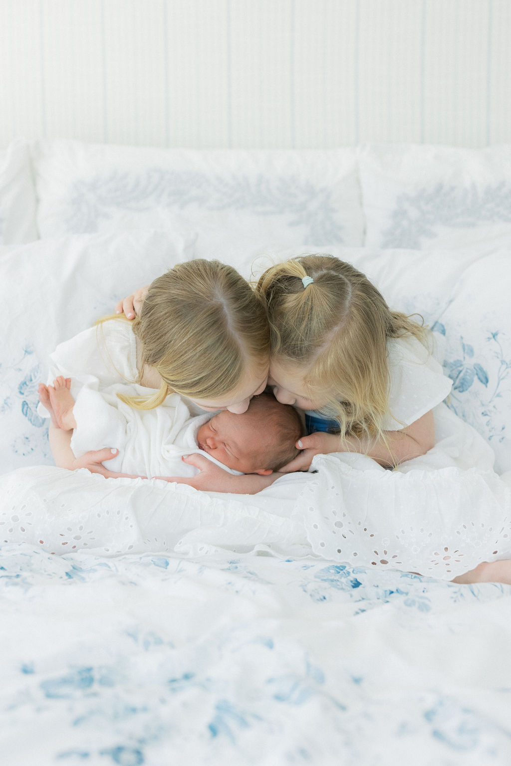 Two young girls sit on a bed in white dresses kissing the head of their newborn baby sister