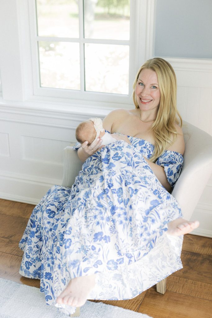 A mother in a blue floral dress sits in a chair under a window with her newborn baby thanks to nannies in new jersey
