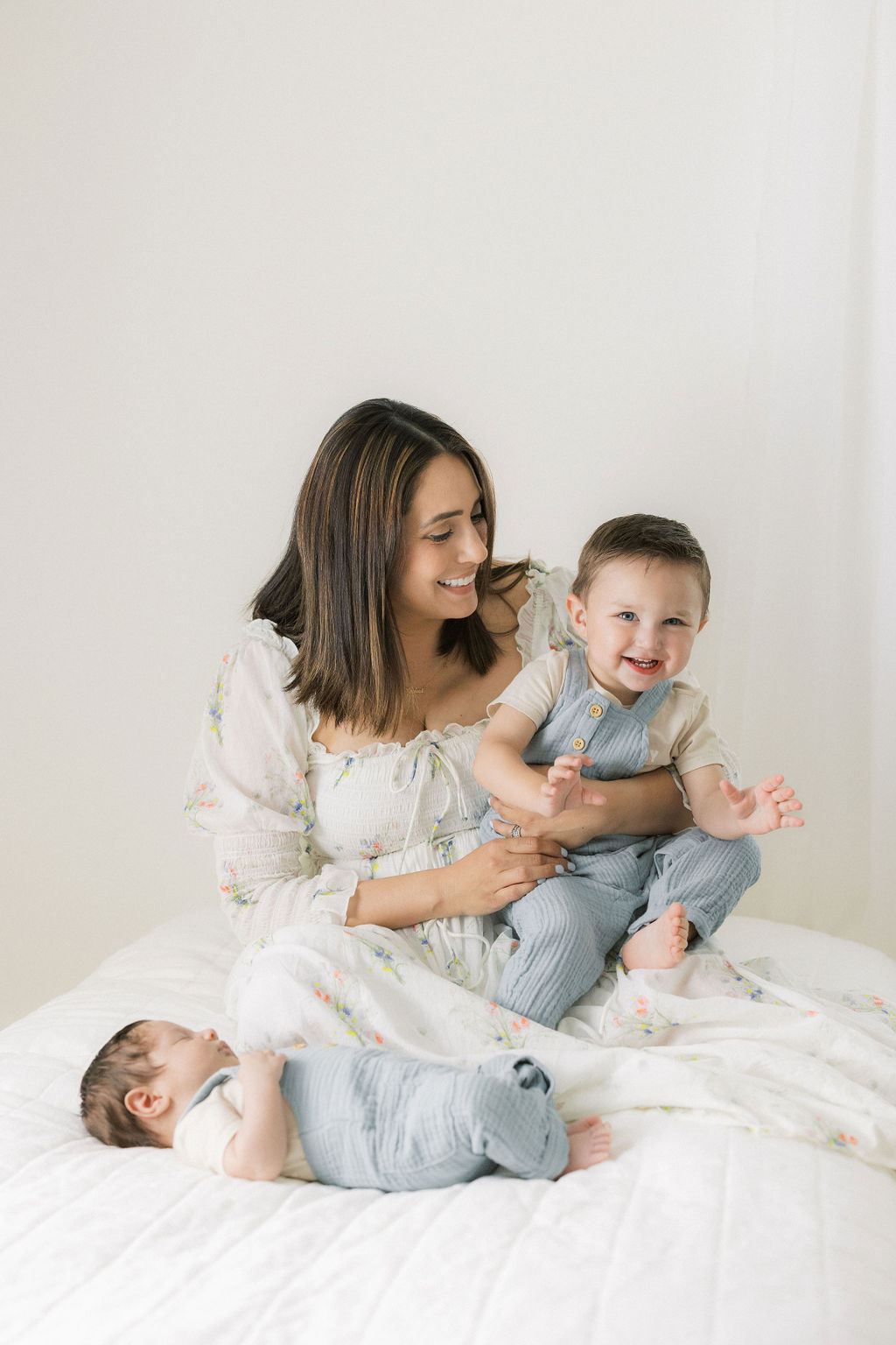 A mother in a dress sits on a bed playing with her toddler son and newborn baby sleeping in front of her after meeting with branchburg lactation