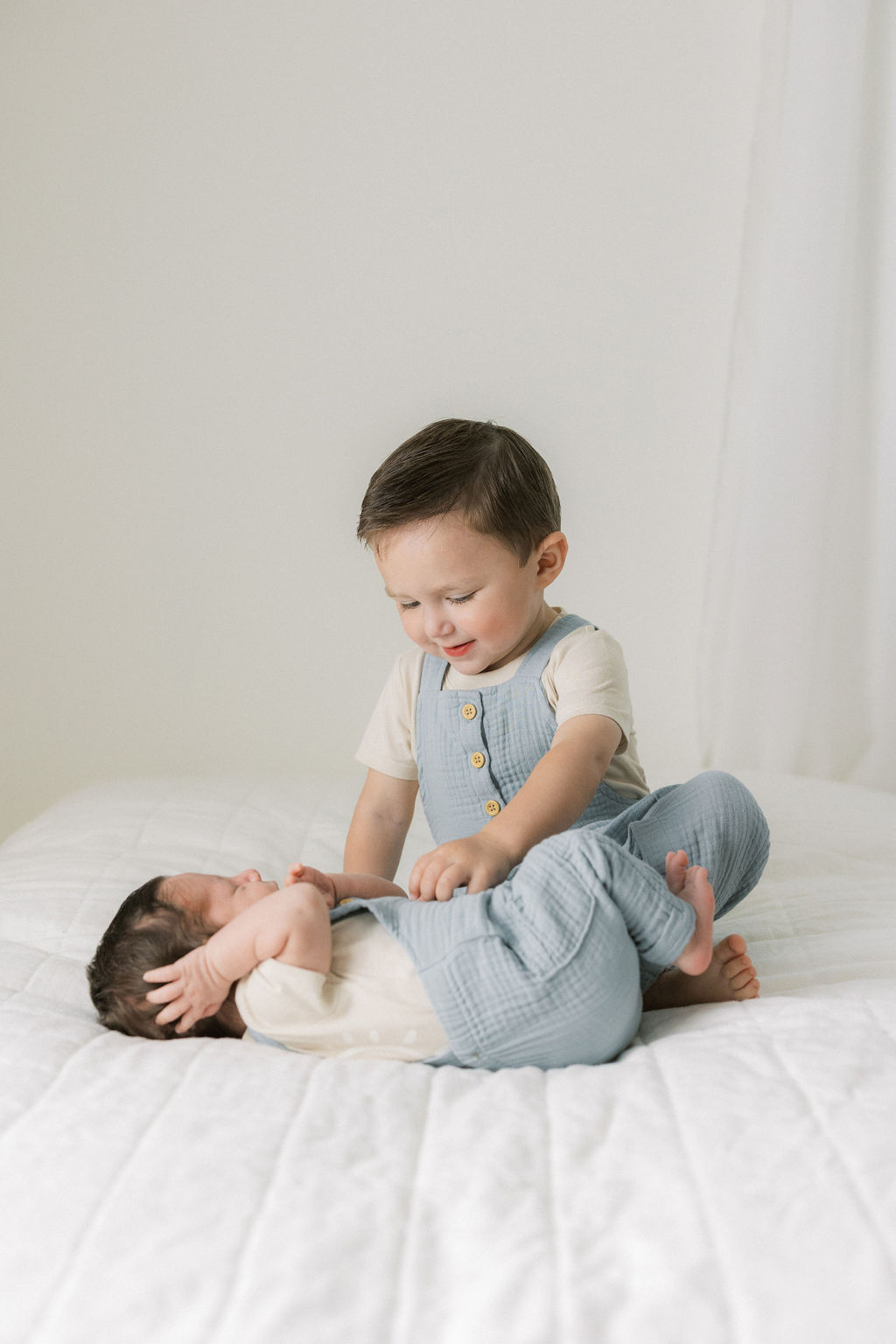 A young toddler boy in blue overalls tickles his newborn baby brother while on a bed in matching overalls after meeting branchburg lactation