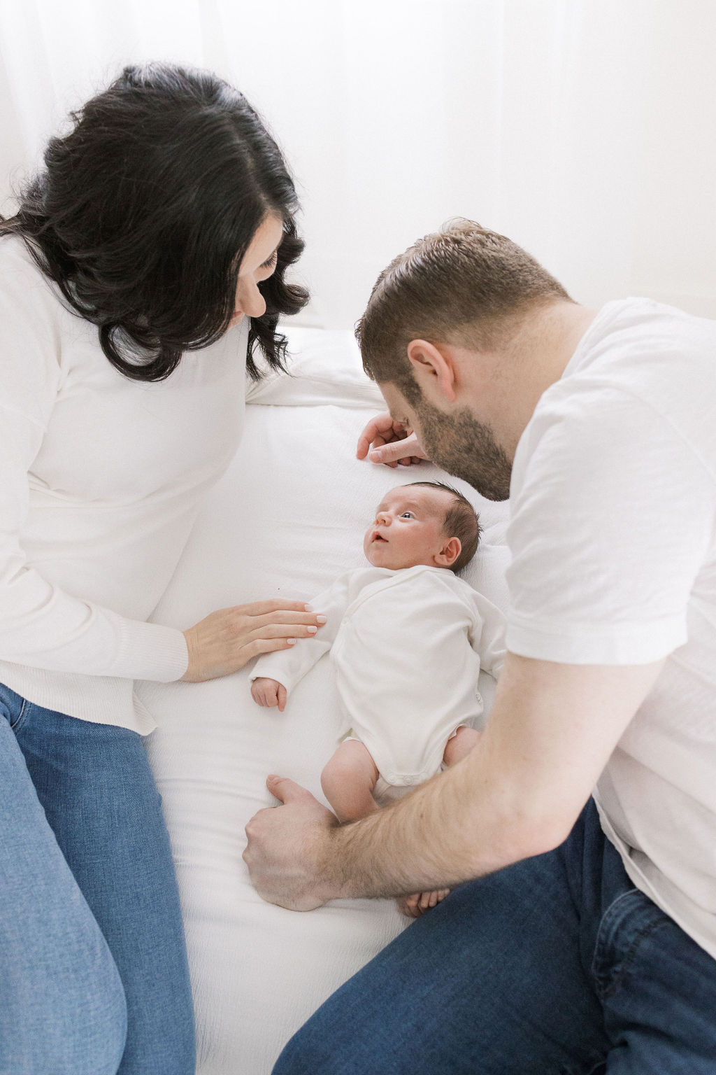 A mom and dad lay with their newborn baby on a bed in a studio in white shirts and jeans