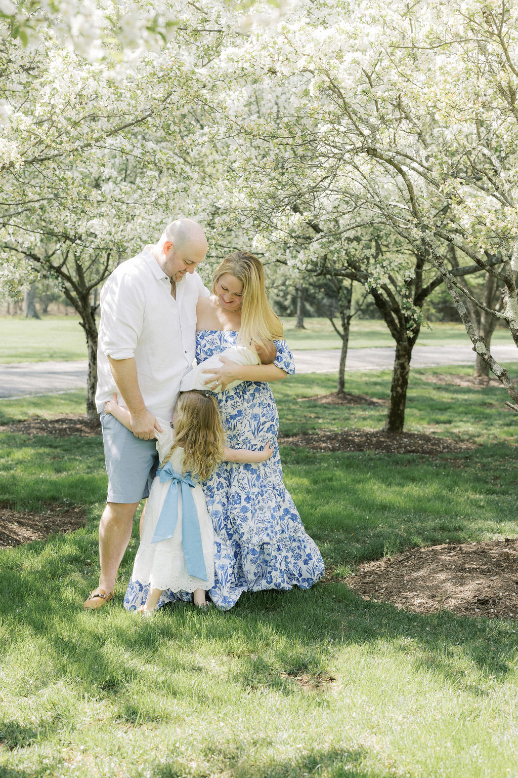 A mom and dad stand in a park holding their newborn baby as their toddler daughter hugs them both after using onecare midwifery morristown