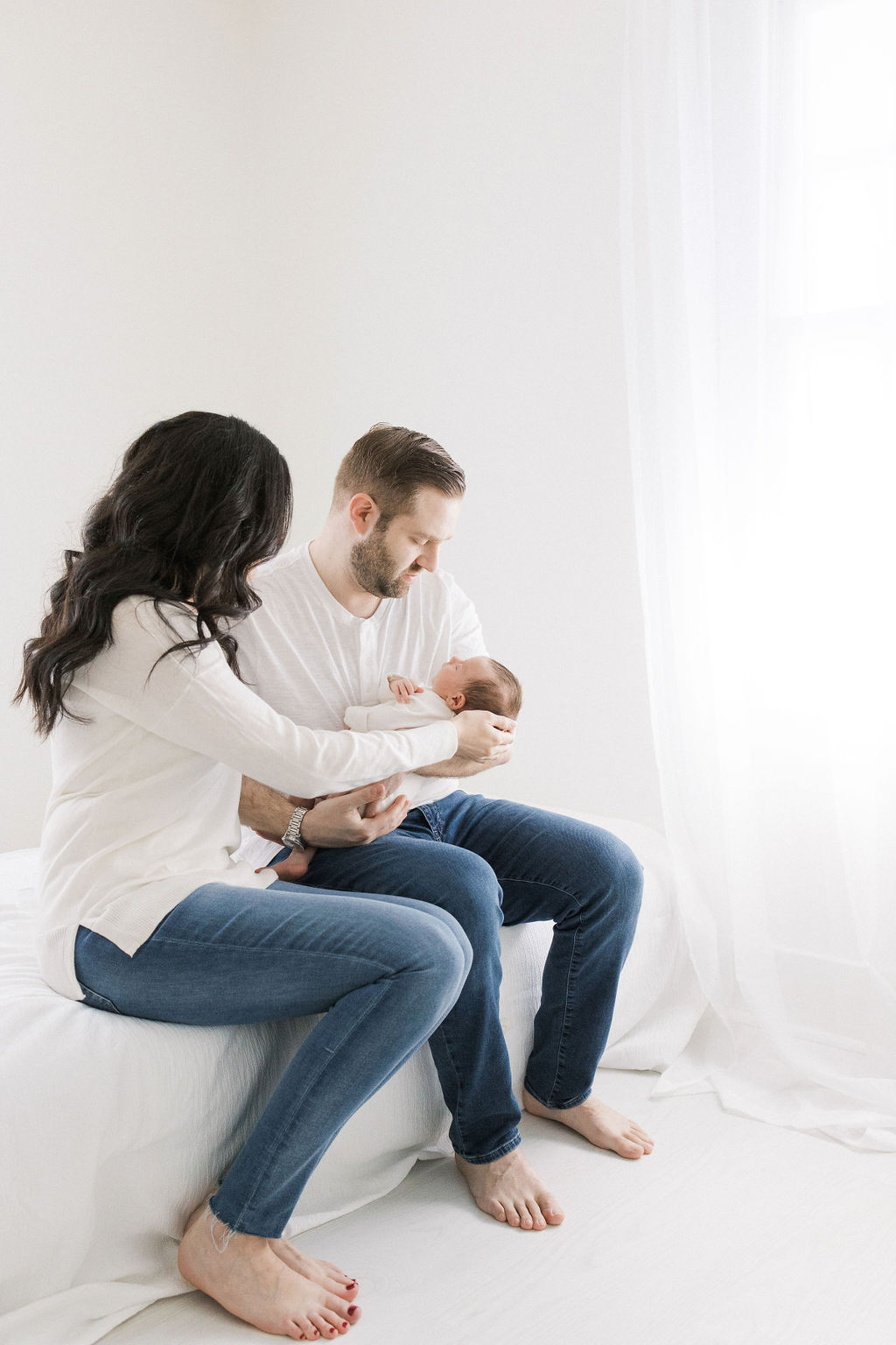 A mom and dad sit on the edge of a bed cradling their sleeping newborn baby in white shirts and jeans after meeting a postpartum doula nj