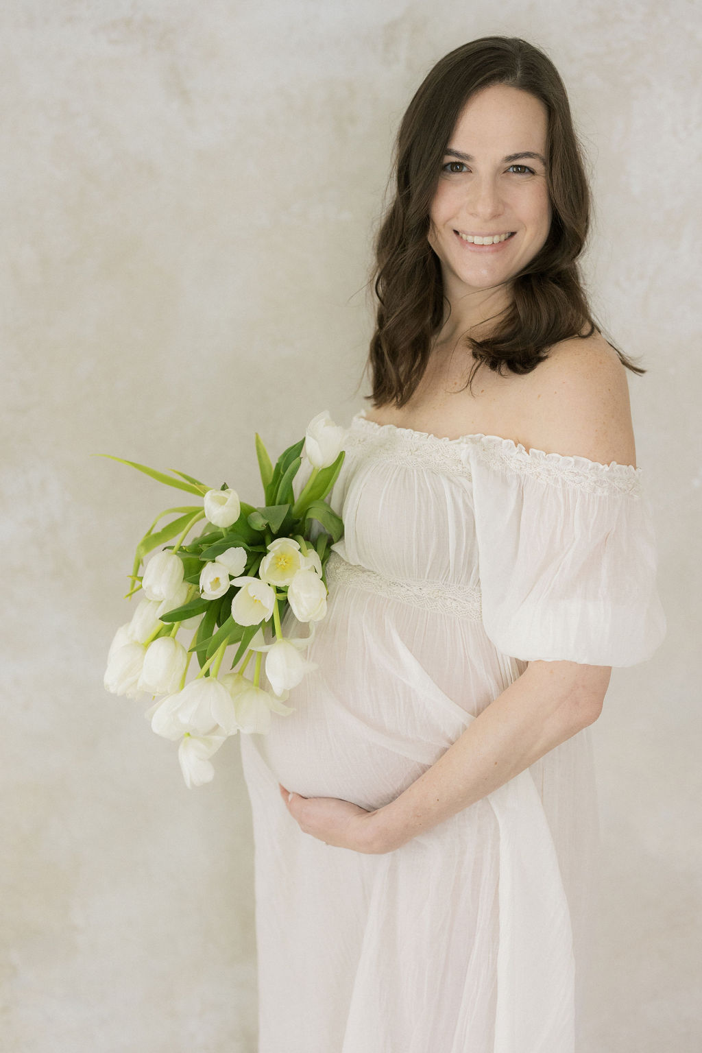 A mother to be stands in a studio holding her bump in a white maternity gown after a 3d ultrasound morristown nj