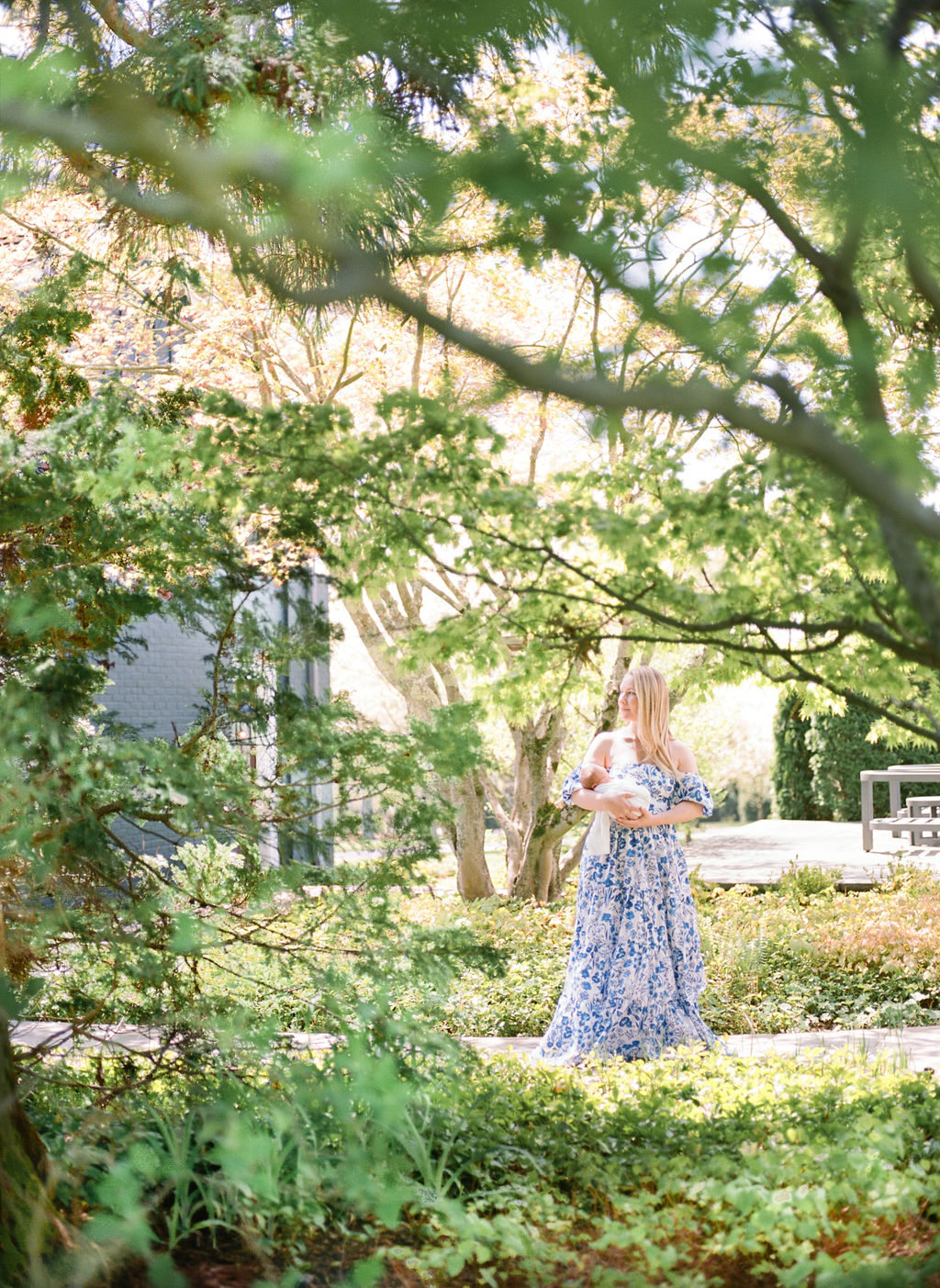 A mother in a blue floral dress stands in a park sidewalk holding her sleeping newborn baby