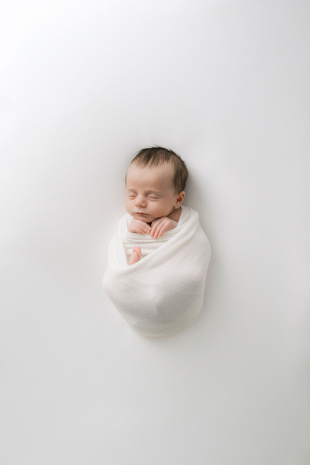 A newborn baby sleeps in a white swaddle with hands sticking out on a white bed