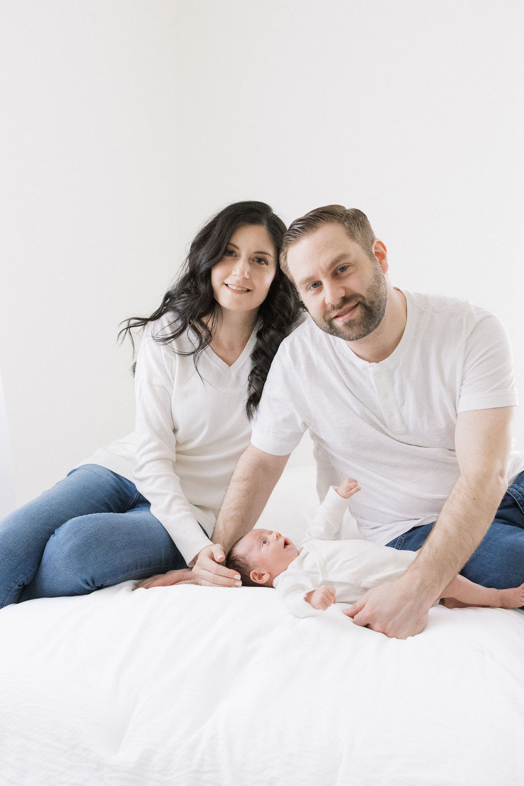 A mother and father in white shirts and jeans sit on a bed over their newborn baby after using a fertility clinic morristown nj