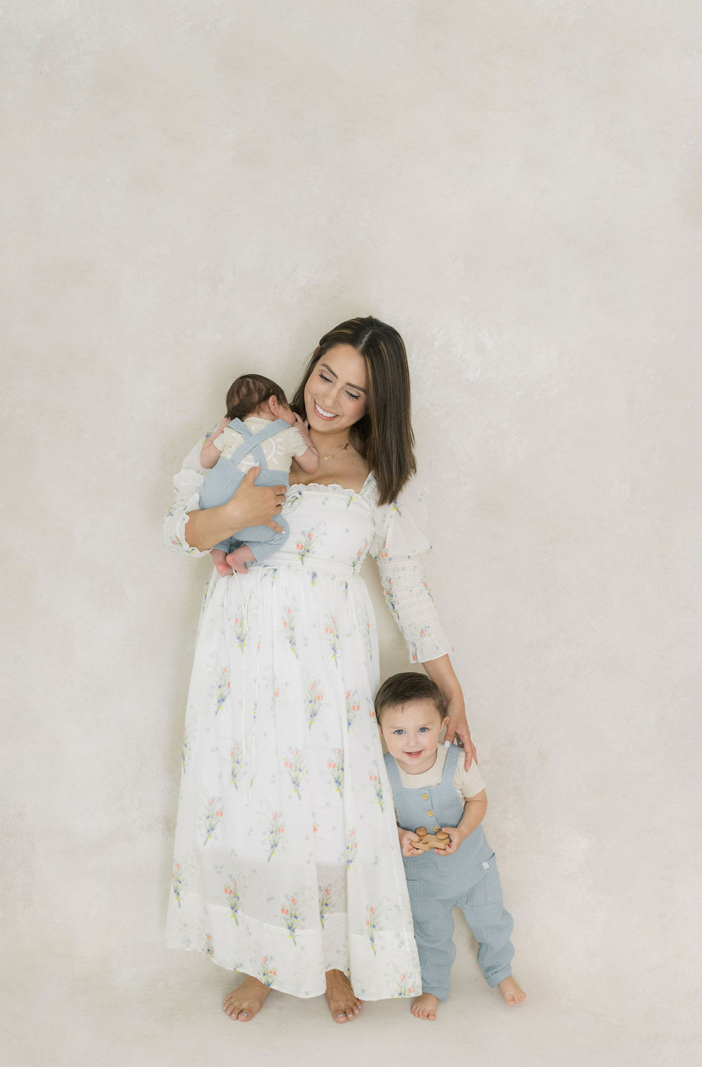 A mother in a white floral print dress stands in a studio holding her newborn baby on her shoulder with her toddler son at her side