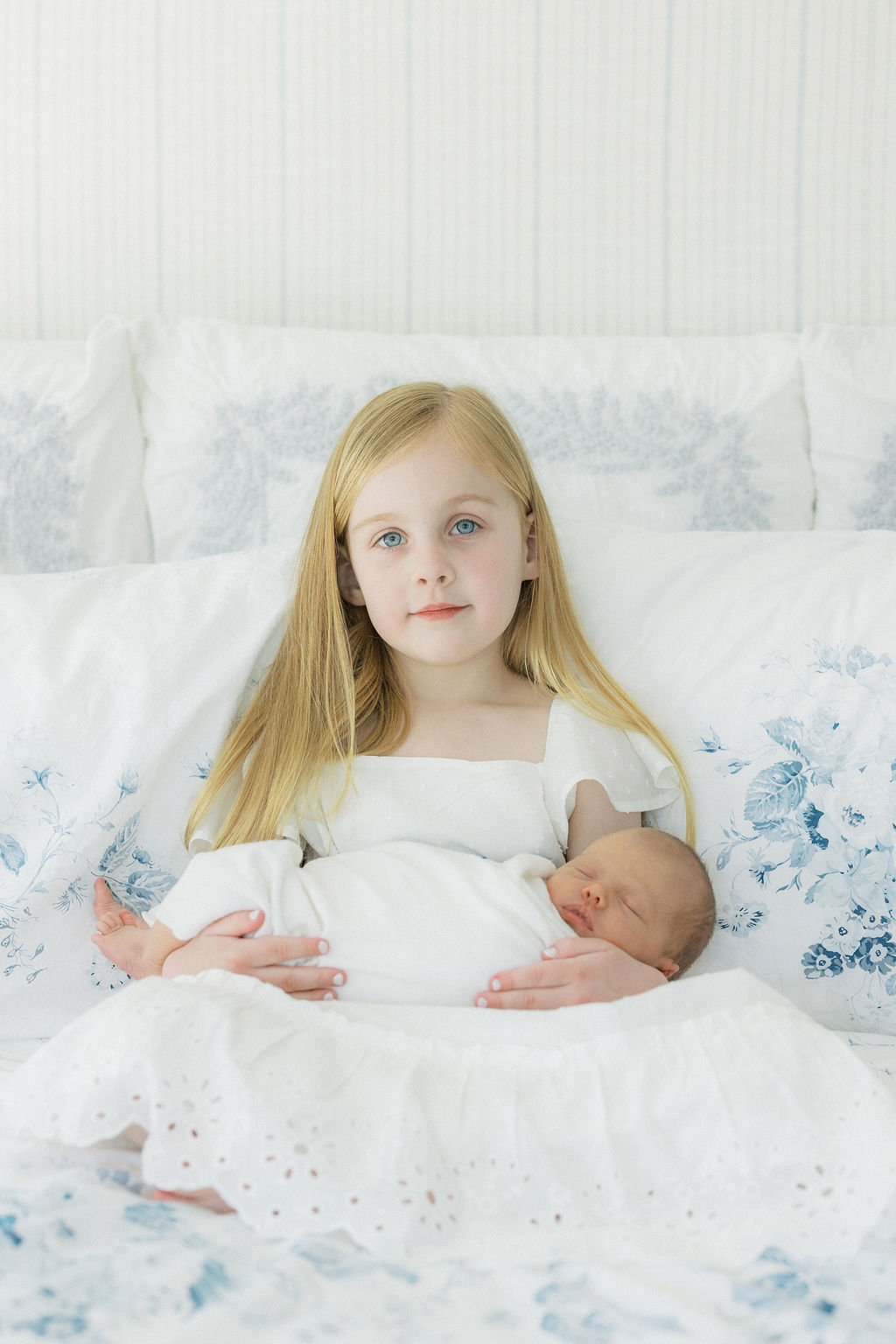 A young girl sits on a floral print bed holding her sleeping newborn baby sibling in her lap
