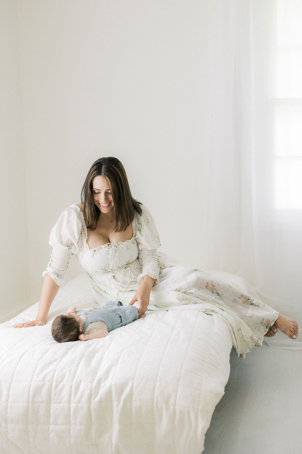 A happy new mother sits on a bed playing with her newborn baby in blue overalls