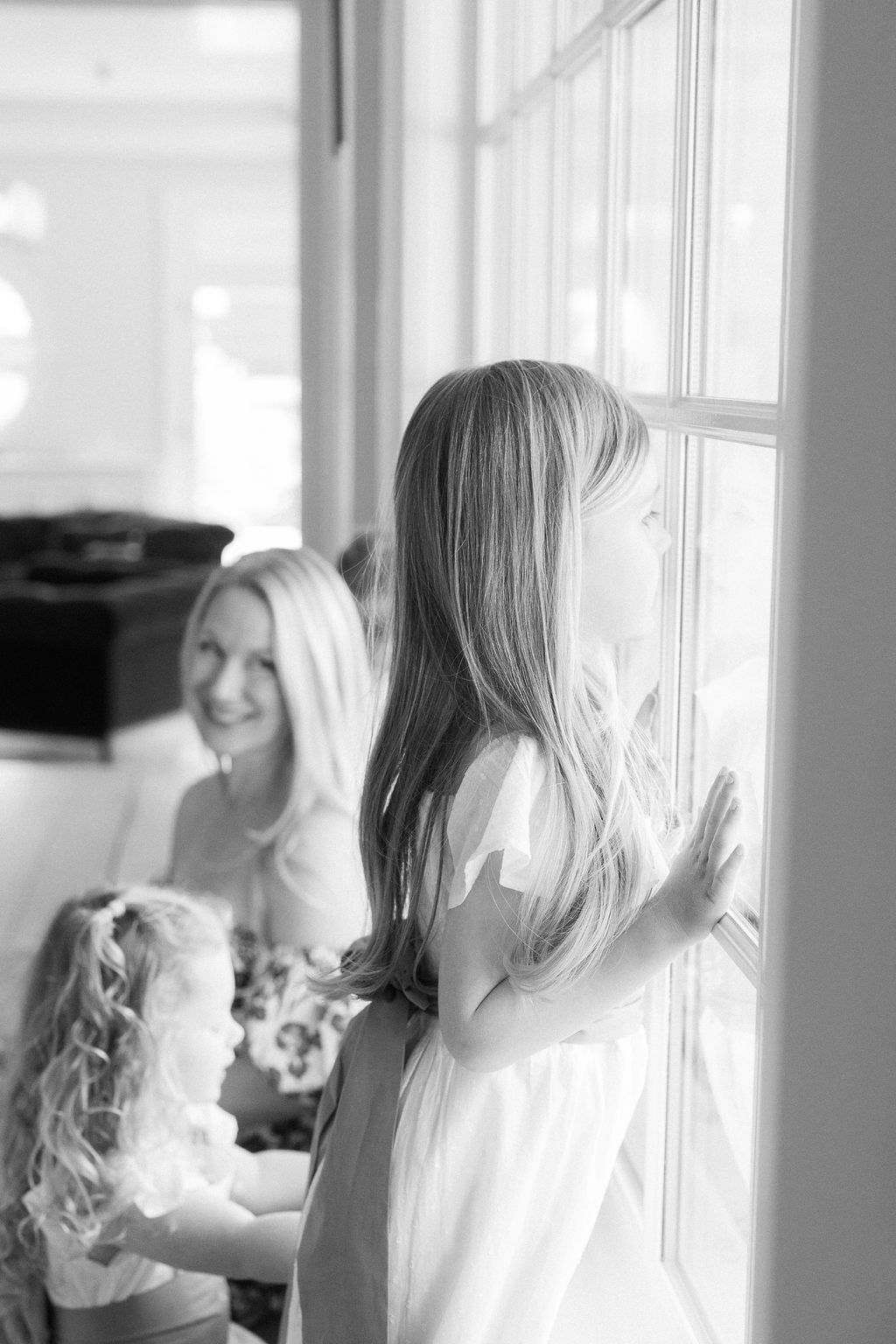 A toddler girl looks out a window while mom and her little sister sit beside her
