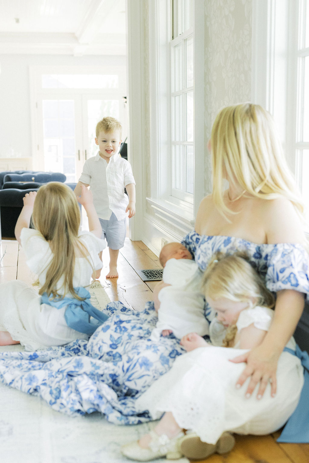 A mother in a blue floral dress plays with her three toddler children while sitting on the floor with her newborn in her arms after a prenatal massage nj