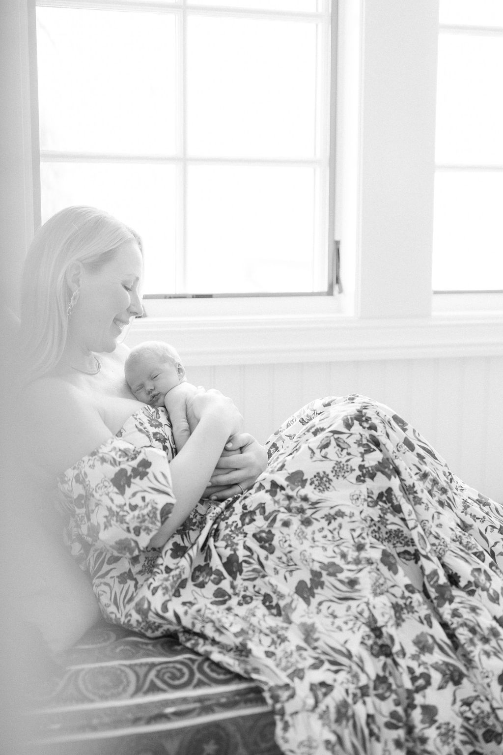 A happy mother in a floral dress sits on a bed in a studio with her newborn baby sleeping on her chest under a window thanks to birthing classes nj