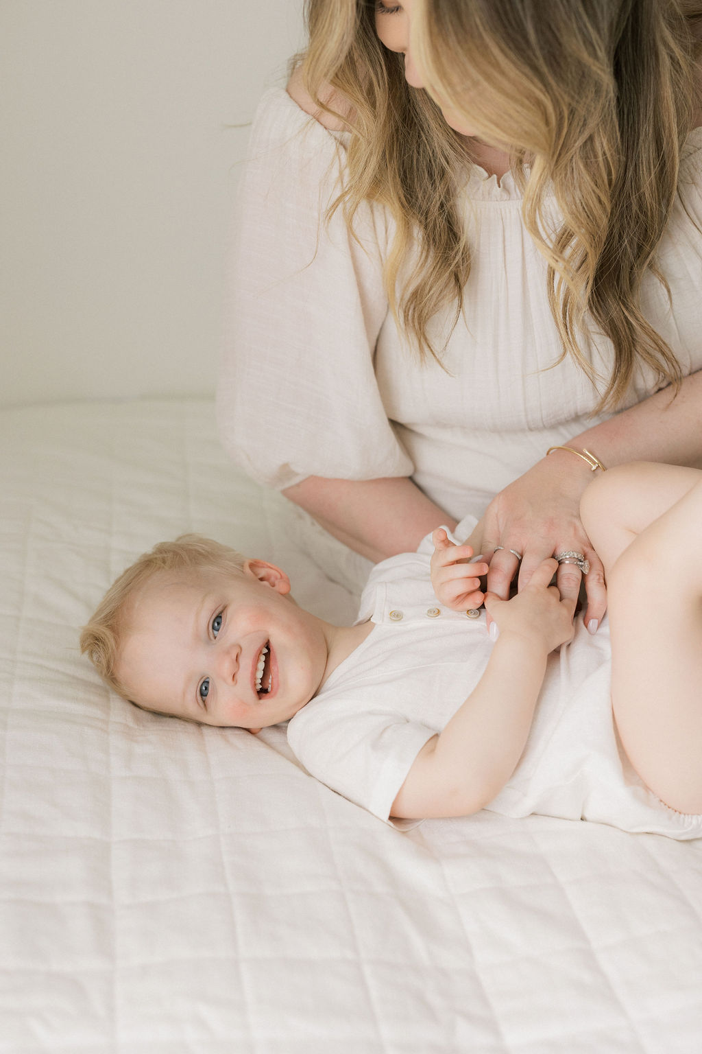 A mother in a white dress sits on a bed tickling her toddler son in a white onesie before meeting a nj au pair
