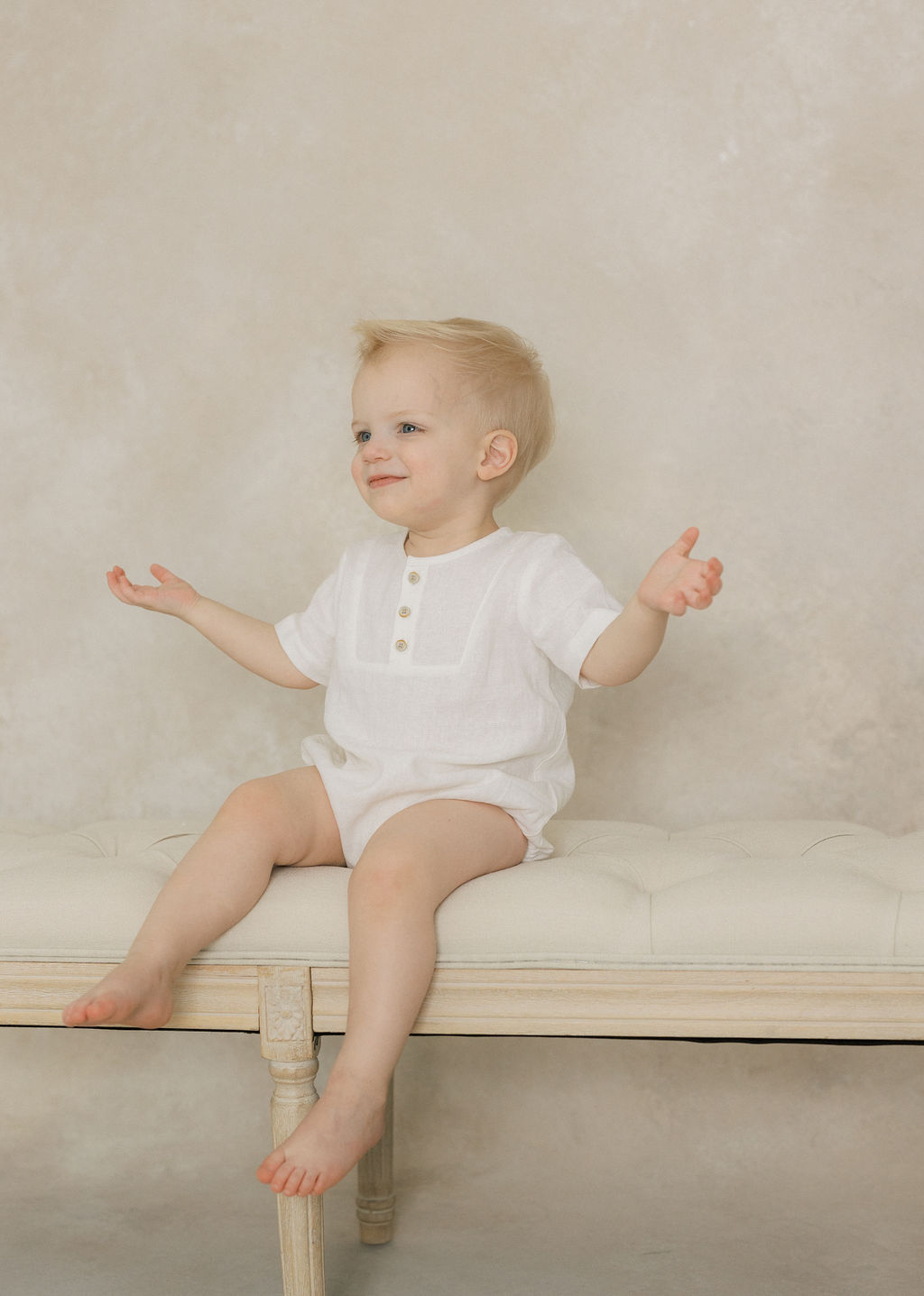 A toddler boy sits on a bench in a studio in a white onesie with arms spread before meeting a nj au pair