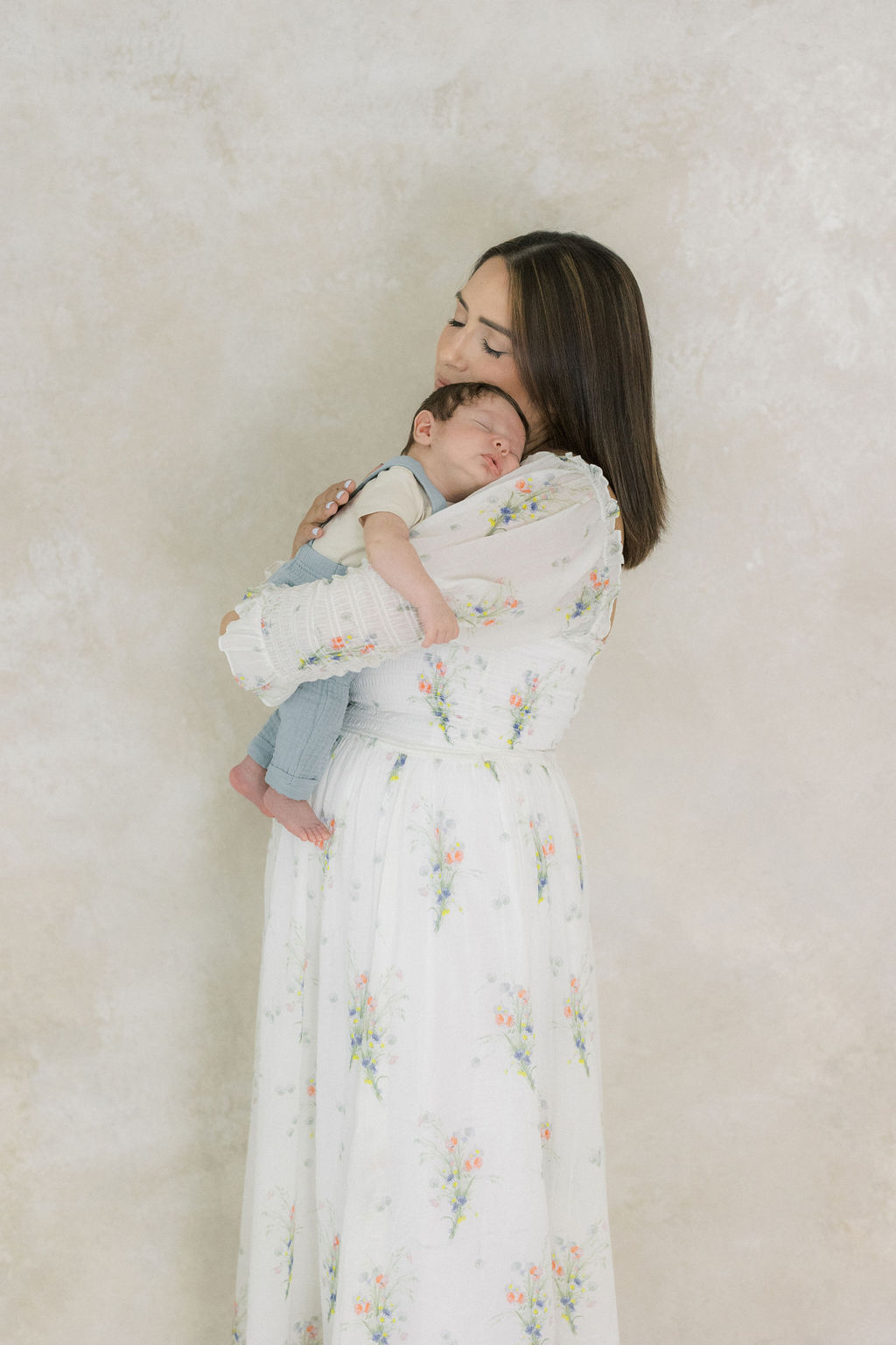A new mother in a white floral print dress cradles her sleeping newborn on her shoulder while standing in a studio after some parenting classes nj