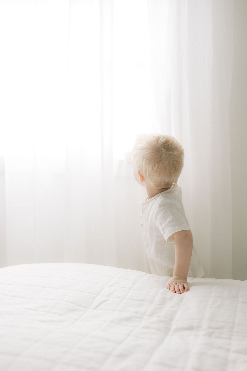 A toddler boy explores a window above a bed in a studio while wearing a white onesie