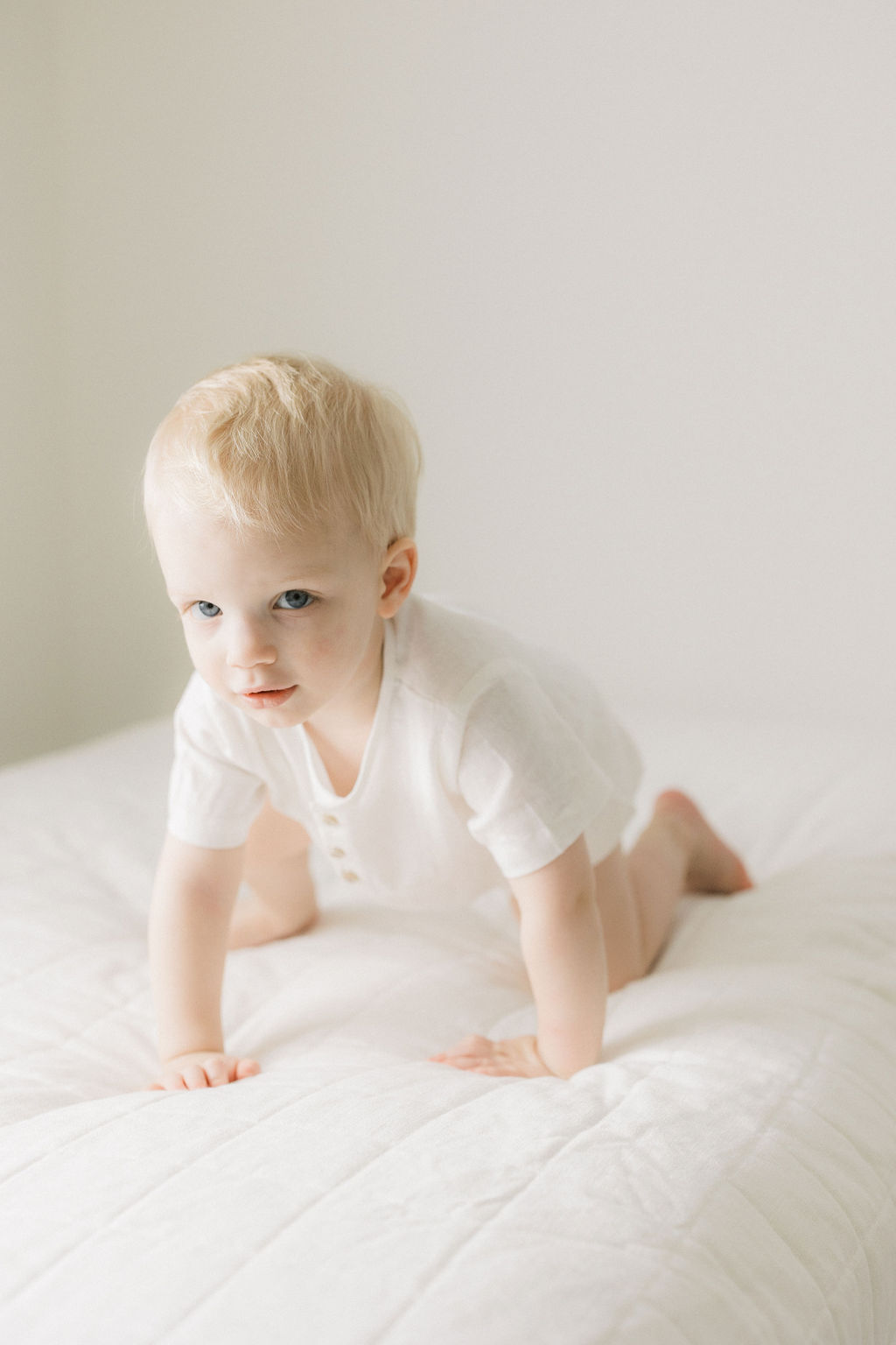A toddler boy in a white onesie crawls on a bed in a studio before meting a pediatric chiropractor nj
