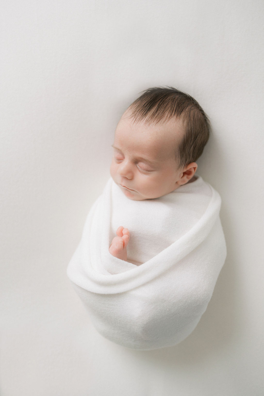 A newborn baby sleeps calmly in a studio while wrapped tight in a white swaddle