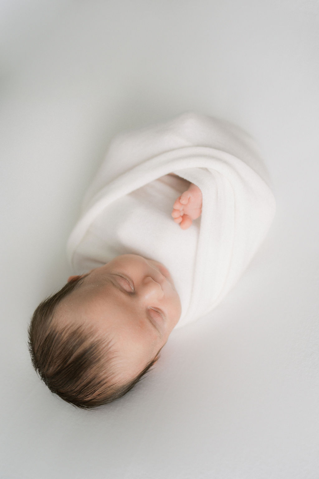 A newborn baby sleeps in a white swaddle with 1 foot sticking out the middle before meeting a pediatric dentist nj