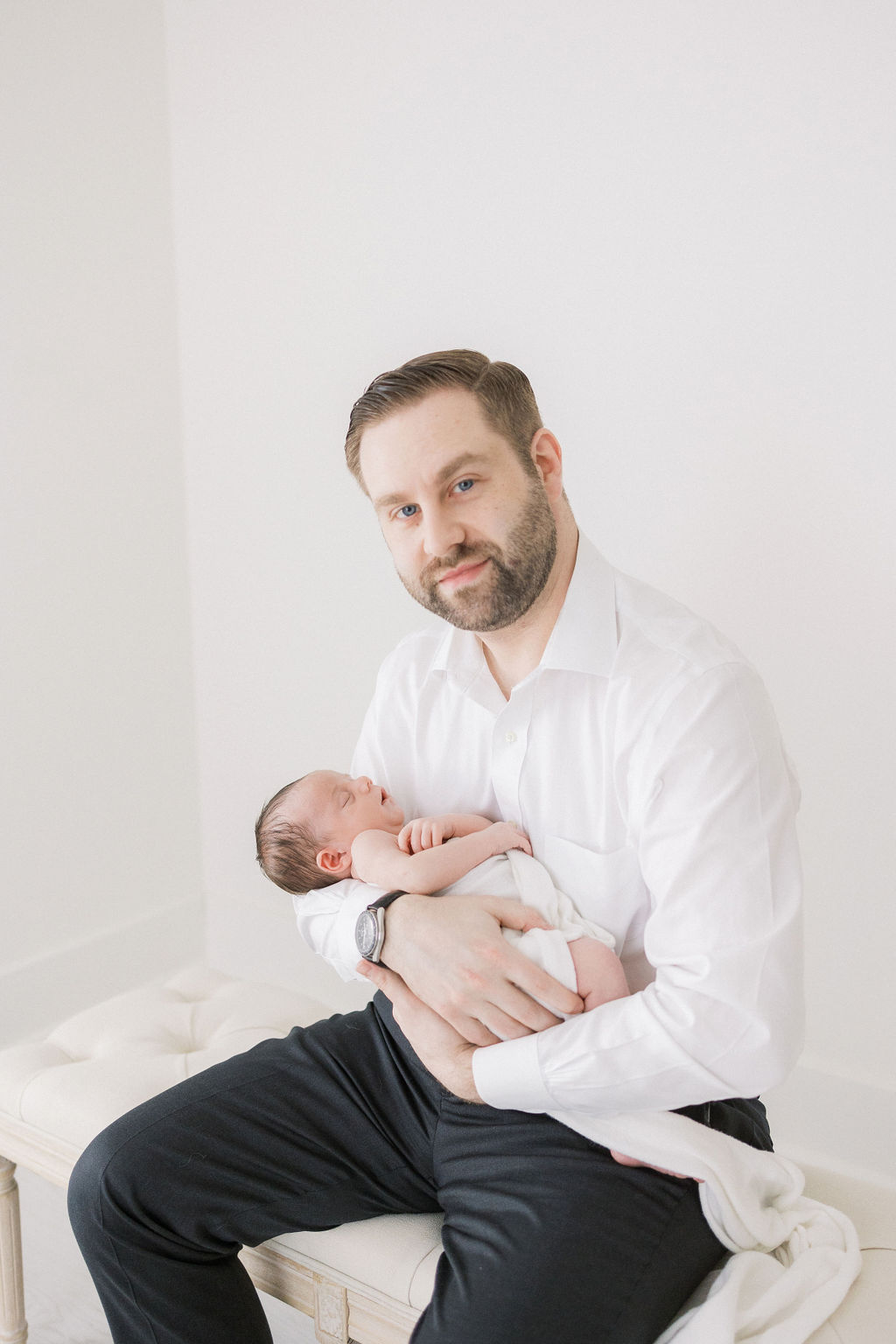 A new father in a white shirt and black pants sits on a bench in a studio cradling his sleeping newborn baby before finding a pediatric dentist nj