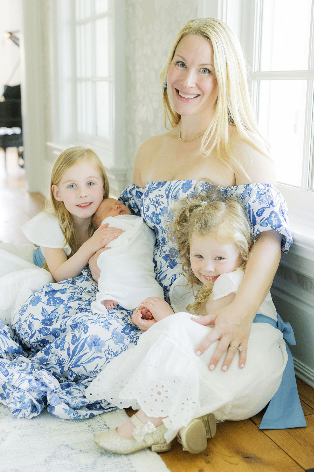 A blonde mother in a blue floral print dress sits on the floor under a window hugging her two toddler daughters in matching white dresses and her newborn baby sleeping in her lap after meeting with a pediatrician nj