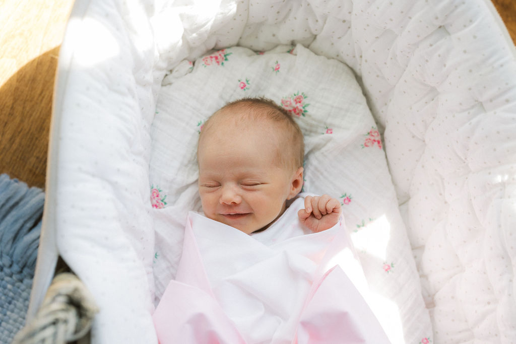 A smiling newborn baby lays in a crib with eyes closed under a pink bow