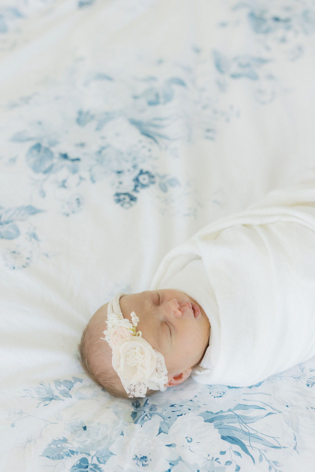 A sleeping newborn baby laying on a bed in a floral headband and white swaddle