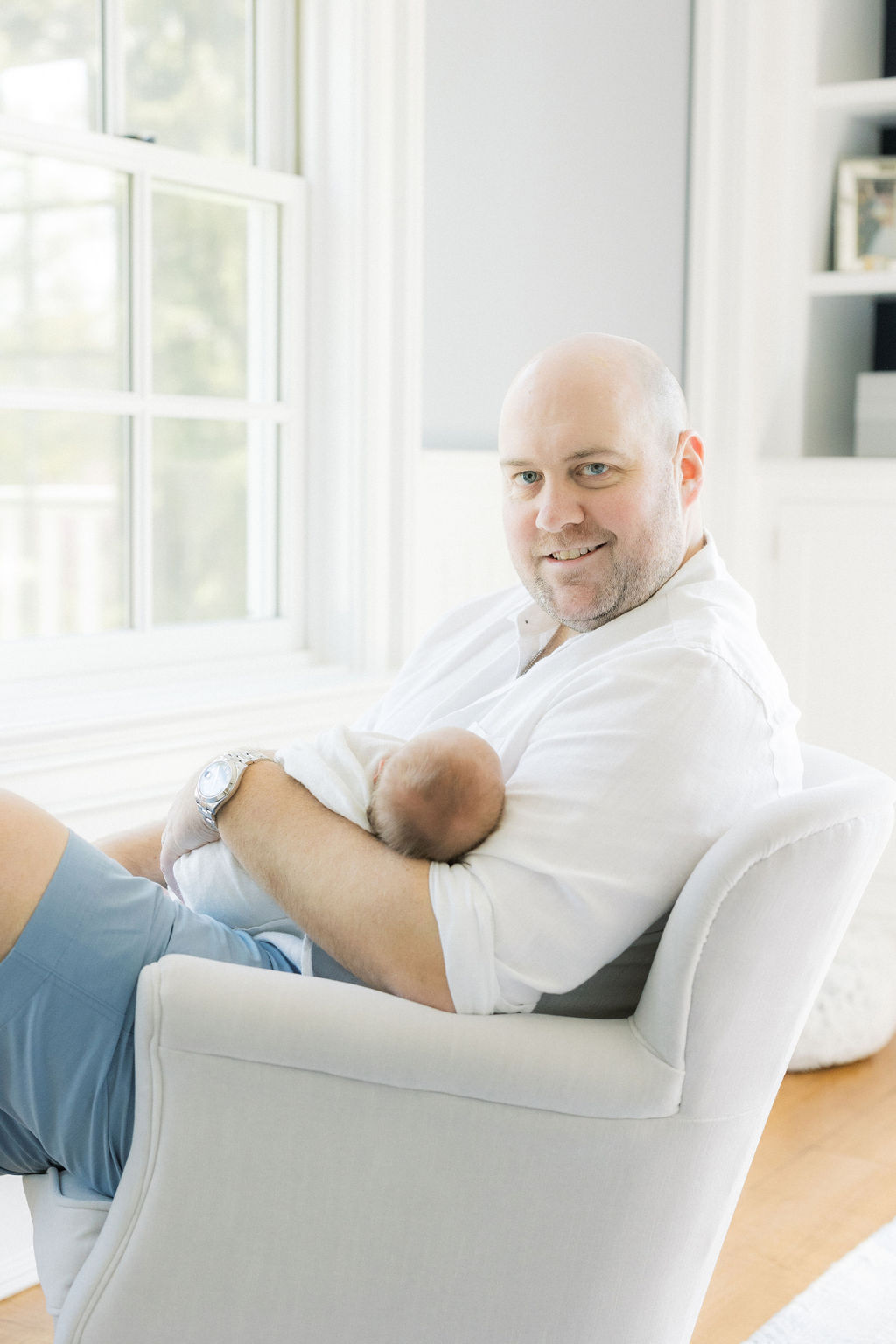 A happy dad sits in a chair smiling with his newborn baby sleeping in his lap