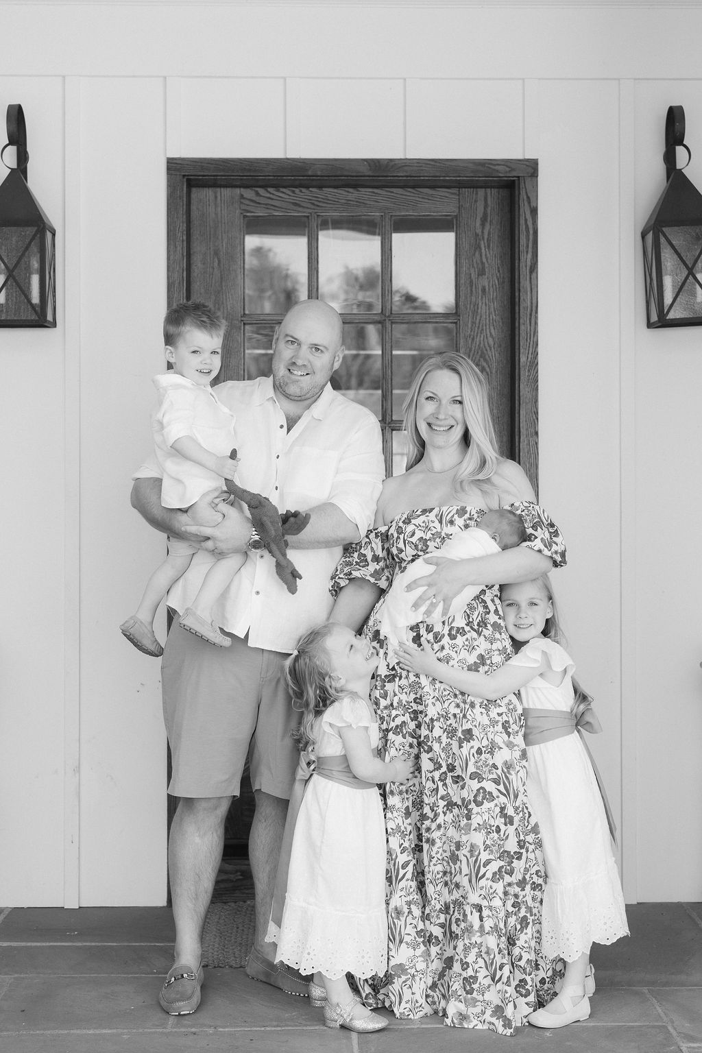 A happy family stands on a porch with two toddler daughters and a toddler son and newborn baby in mom's arms after using placenta encapsulation nj